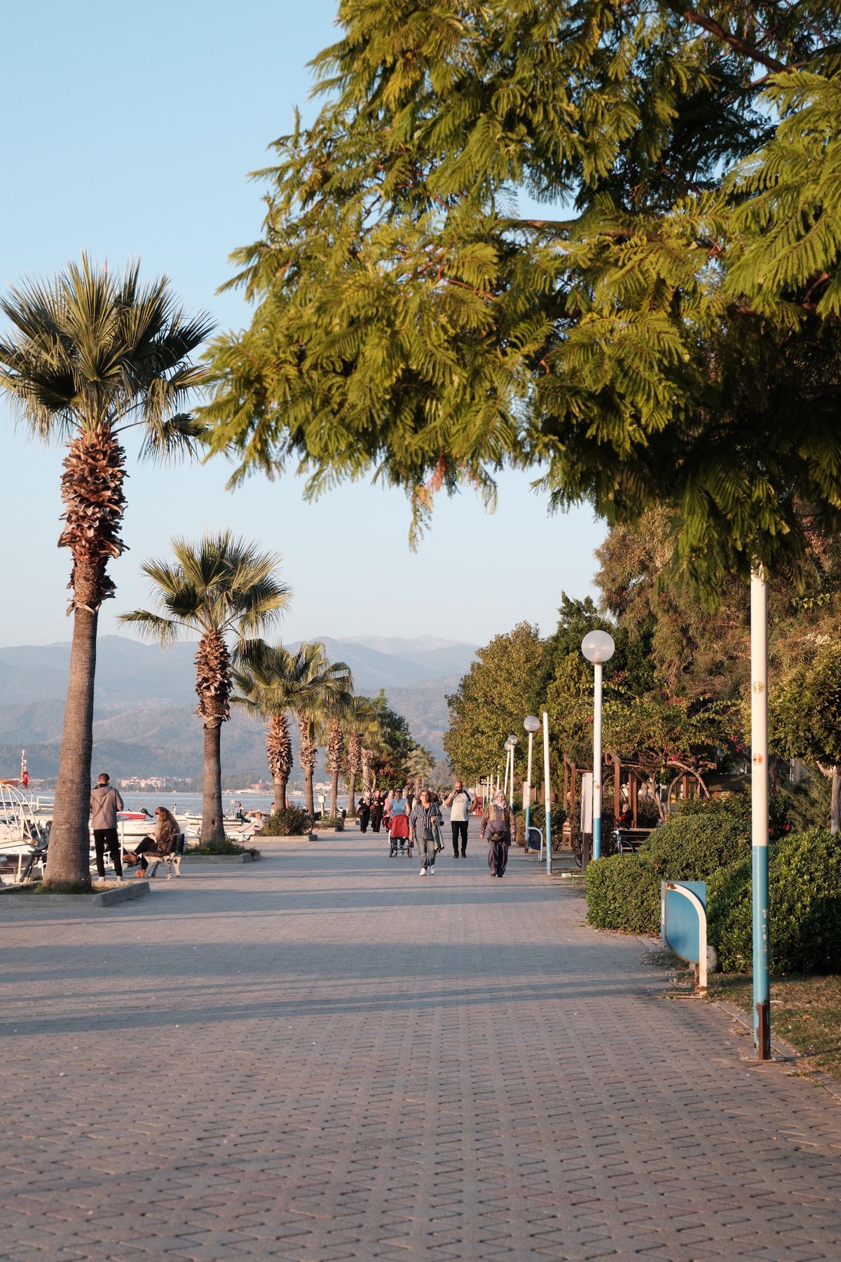 Palm-lined seafront promenade at Fethiye in evening light with people walking, moored boats to one side and hazy mountains across the bay